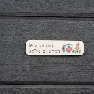 May include: A wooden sign with the text "Je vide ma boîte à lunch" in black lettering. The sign also features a cartoon illustration of a lunchbox, an apple, a juice box, and a small container.