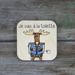 Puede incluir: Un letrero de madera con un alce de dibujos animados sentado en un inodoro y leyendo un libro. El letrero dice "Je vais à la toilette" en francés.