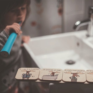 May include: Four wooden coasters with illustrations of a moose. Each coaster has a different French phrase printed on it: "Je vais a la toilette", "Je prends un bain", "Je mets mon pyjama", and "Je brosse mes dents".