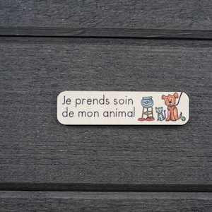 K&ouml;nnte beinhalten: Ein wei&szlig;es Holzschild mit dem Text "Je prends soin de mon animal" in schwarzen Buchstaben. Das Schild zeigt eine Comic-Illustration eines Hundes, einer Katze und einer Fischschale.