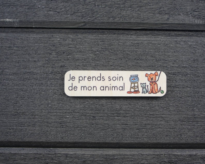 K&ouml;nnte beinhalten: Ein wei&szlig;es Holzschild mit dem Text "Je prends soin de mon animal" in schwarzen Buchstaben. Das Schild zeigt eine Comic-Illustration eines Hundes, einer Katze und einer Fischschale.