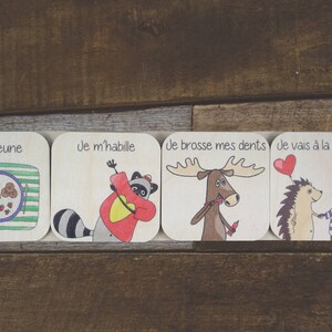 May include: Four wooden cards with illustrations and French text. The cards show a raccoon eating breakfast, a raccoon getting dressed, a moose brushing its teeth, and a hedgehog and a bear going to daycare. The text on the cards reads "Je déjeune", "Je m'habille", "Je brosse mes dents", and "Je vais à la garderie".