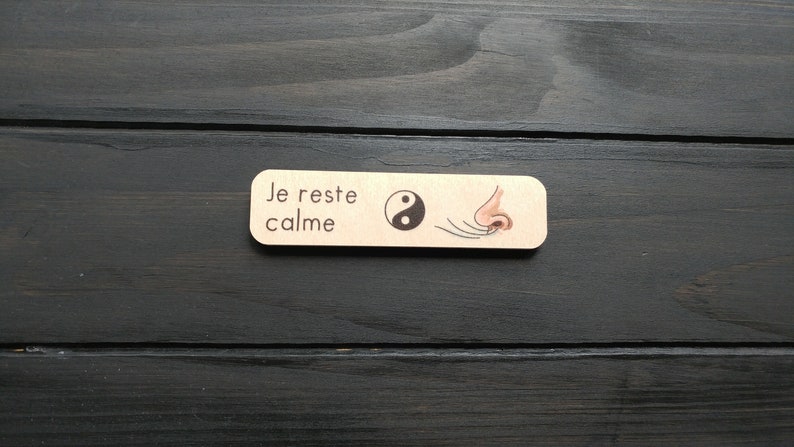 May include: A wooden bookmark with the text "Je reste calme" and a black and white yin yang symbol. The bookmark also features a drawing of a nose with lines coming out of the nostrils.