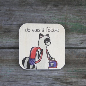 Puede incluir: Un letrero de madera con un mapache de dibujos animados que lleva una mochila y una fiambrera. El letrero dice "Je vais à l'école", que en francés significa "Voy a la escuela."