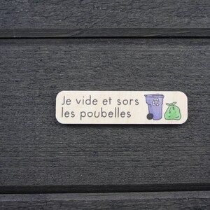 Puede incluir: Un letrero de madera blanco con el texto "Je vide et sors les poubelles" e ilustraciones de un contenedor de reciclaje morado y una bolsa de basura verde.
