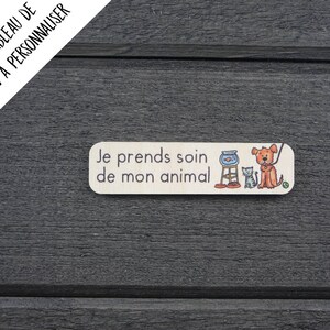 K&ouml;nnte beinhalten: Ein rechteckiges Holzschild mit dem Text "Je prends soin de mon animal" in schwarzer Schrift. Das Schild zeigt eine Comic-Illustration eines Hundes, einer Katze und einer Fischschale.