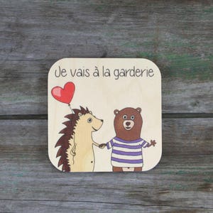 Puede incluir: Una tarjeta de madera con un erizo y un oso cogidos de la mano. La tarjeta tiene el texto "Je vais à la garderie" escrito en ella. El erizo lleva un globo rojo en forma de corazón.