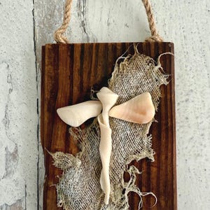 May include: A wooden wall hanging with a natural, rustic aesthetic. It features a dark brown, rectangular wooden base with a jute rope handle. A cross-shaped design is crafted from seashells and burlap.