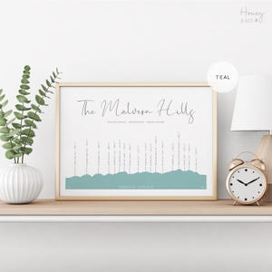 Op de afbeelding: Een ingelijste horizontale print getiteld The Malvern Hills in een blauwgroen kleurenpalet. Het kunstwerk toont een gestileerd silhouet van een bergketen met verticale lijnen die wijzen naar specifieke bergnamen en hoogtes in voet op een witte achtergrond.
