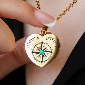 Puede incluir: Un collar con colgante en forma de corazón dorado. El colgante presenta un diseño de brújula con las coordenadas "52°31'12" N 13°24'18" E". La cadena es fina.