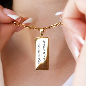 Puede incluir: Un collar con colgante rectangular dorado con el texto "48.8566° N, 2.3522° E Our Forever Place". El collar lo sostiene una persona que lleva una camisa blanca.