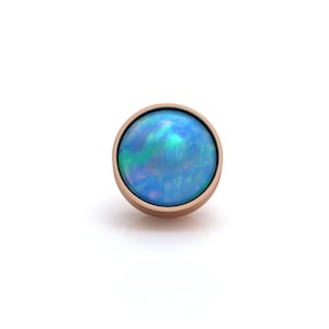 May include: A circular stud earring featuring a smooth, cabochon-cut blue opal gemstone with iridescent green and violet flashes, set within a polished rose gold metal bezel. The design is simple and round, highlighting the vibrant play of color.