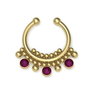 Puede incluir: Un anillo de septum dorado con un diseño de aro circular adornado con una serie de pequeñas esferas metálicas y tres piedras preciosas redondas de color magenta intenso dispuestas a lo largo del borde inferior de la pieza.