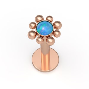 May include: A rose gold flat back labret stud featuring a flower-shaped top. The center of the flower is a round, iridescent blue opal cabochon, surrounded by eight small, smooth rose gold spheres arranged in a circular petal pattern.