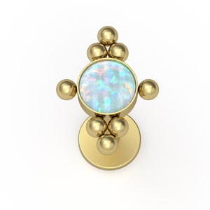 May include: A gold-toned flat back stud earring featuring a central circular iridescent opal cabochon. The setting is accented with small gold spheres arranged in a cross-like pattern above, below, and on the sides of the central stone.