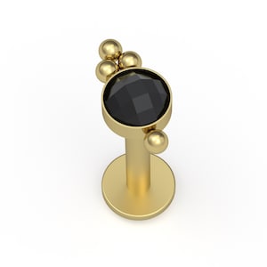 May include: A gold-toned labret stud featuring a circular setting with a faceted black stone. The top of the setting is adorned with three small gold spheres, and a single gold sphere is positioned on the side of the circular stone housing.