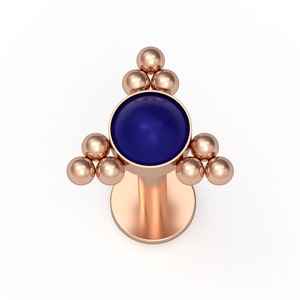 May include: A rose gold flat back stud earring featuring a central circular deep blue stone. The setting is accented by three small metallic spheres arranged in a triangular cluster on the left, right, and top sides of the central stone.