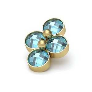 May include: A gold-toned jewelry piece featuring four round, faceted light blue stones arranged in a cluster. Two small, smooth gold spheres are positioned in the center between the stones, creating a flower-like shape with a metallic setting.