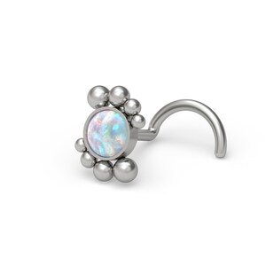 Op de afbeelding: Een zilverkleurige neuspiercing met een centrale ronde iriserende opaalsteen, omringd door een cluster van kleine gepolijste metalen bolletjes in een halvemaanvorm, bevestigd aan een gebogen staafje voor een veilige plaatsing in de piercing.