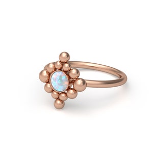May include: A rose gold ring featuring a central round opal cabochon surrounded by a cluster of small, smooth metal spheres arranged in a diamond-shaped pattern. The band is thin and polished, matching the rose gold tone of the setting.