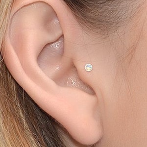 May include: A close-up of a person's ear with a small opal stud earring. The earring is a simple, minimalist design and is set in a silver or white gold setting.