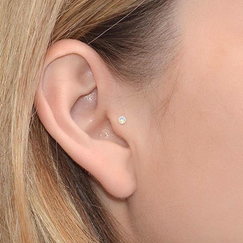 May include: A close-up of a person's ear with a small opal stud earring. The earring is a simple, minimalist design and is set in a silver or white gold setting.