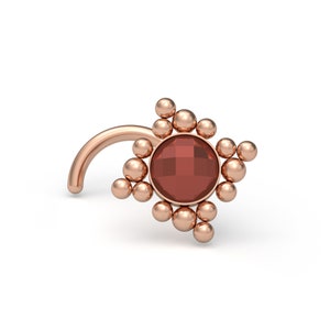 Peut inclure: Un bijou de nez en or rose avec une pierre précieuse ronde à facettes rouge foncé au centre. La pierre est entourée d'un groupe de petites sphères lisses en or rose disposées en motif géométrique, fixées sur une tige incurvée en or rose.