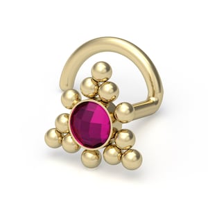 Puede incluir: Un anillo de nariz de oro con una piedra preciosa central circular facetada de color rosa oscuro. La piedra está rodeada por una disposición decorativa de pequeñas esferas doradas en un patrón de racimo triangular, unida a un poste curvo.