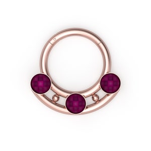 May include: A rose gold septum ring with three round, dark pink gemstones. The ring is a hoop with a slightly thicker band on the top and bottom.