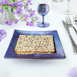 May include: A blue and purple square plate with a stack of matzah on top. A glass of wine with the Hebrew words "L'Chaim" written on it is in the background.