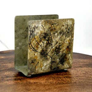 May include: A square-shaped napkin holder with a green, brown, and gold abstract design featuring a tree with branches and small gold dots. The holder has a slightly curved base.
