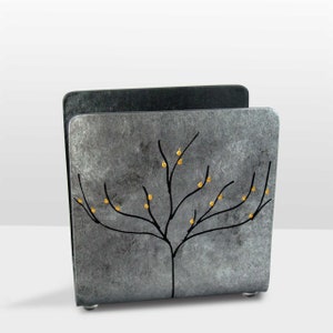 May include: A silver metal napkin holder with a black tree design and gold accents. The holder has a textured surface and a square shape.