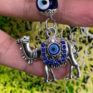 May include: A silver camel charm with a blue evil eye charm on its back. The evil eye charm is surrounded by rhinestones. The charm is attached to a silver chain with a blue evil eye bead.