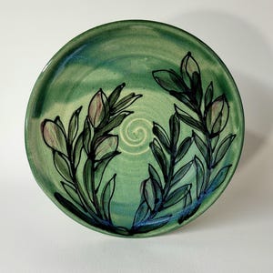 May include: A handmade ceramic plate with a green glaze. The plate features a hand-painted floral design with green leaves and pinkish-purple buds. A spiral design is in the center of the plate.