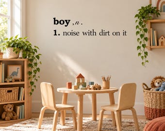 May include: A wall decal featuring the black text 'boy, n. 1. noise with dirt on it' displayed on a plain wall above a wooden children's table and two chairs, surrounded by potted plants, a bookshelf, and a woven basket with toys.