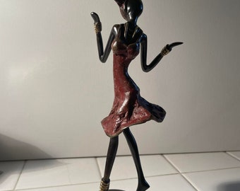 May include: A black and red stylised sculpture of a woman in a dress with a head wrap. The sculpture is on a black and gold base.
