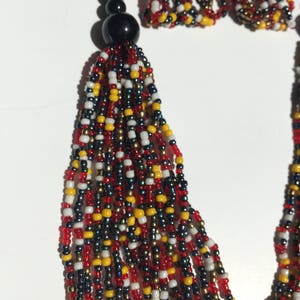 May include: A multi-strand beaded necklace and earrings set. The necklace features a black tassel and is made with red, white, black, and gold beads. The earrings are small and match the necklace.