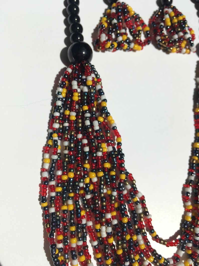 May include: A multi-strand beaded necklace and earrings set. The necklace features a black tassel and is made with red, white, black, and gold beads. The earrings are small and match the necklace.