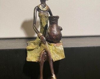 May include: A bronze figurine of a woman wearing a green dress and a head wrap, holding a brown pot. The figurine is sitting on a ledge.