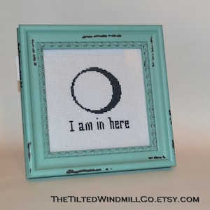 May include: A framed cross-stitch with a black circle and the text "I am in here". The frame is a light teal colour with black distressed edges.