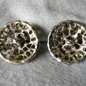 May include: Two silver-toned, filigree-style buttons with a circular design. The buttons have a detailed, openwork pattern and a slightly tarnished appearance, suggesting an antique or vintage aesthetic. They rest on a gray fabric surface.