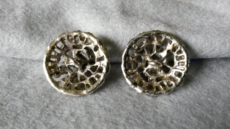 May include: Two silver-toned, filigree-style buttons with a circular design. The buttons have a detailed, openwork pattern and a slightly tarnished appearance, suggesting an antique or vintage aesthetic. They rest on a gray fabric surface.