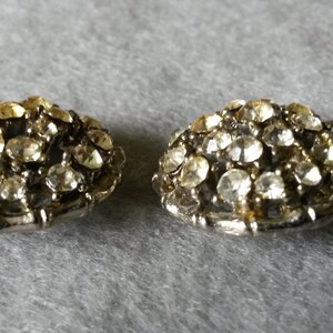 May include: Two vintage, silver-toned, dome-shaped earrings. Each earring is encrusted with numerous clear, sparkling rhinestones. The earrings are displayed on a gray surface, showcasing their intricate design and vintage appeal.