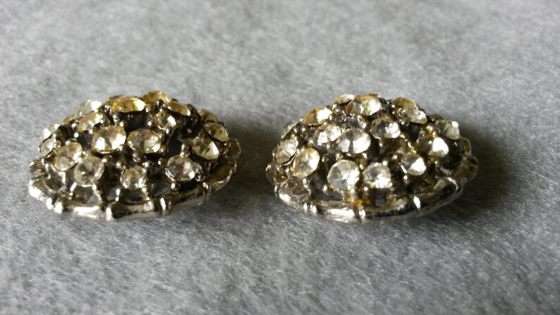 May include: Two vintage, silver-toned, dome-shaped earrings. Each earring is encrusted with numerous clear, sparkling rhinestones. The earrings are displayed on a gray surface, showcasing their intricate design and vintage appeal.