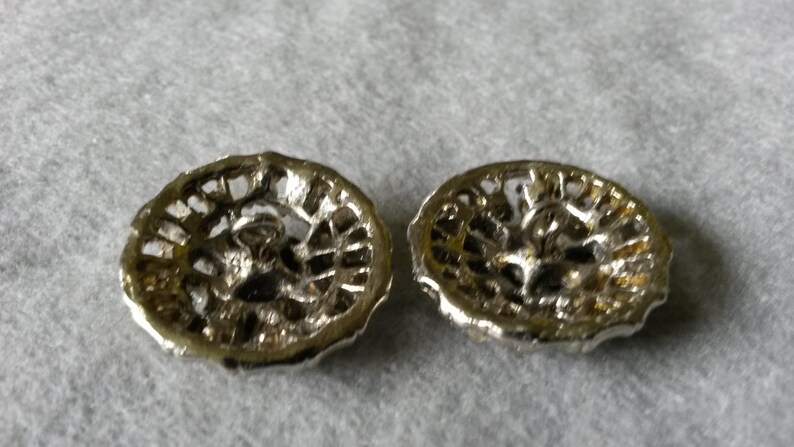 May include: Two silver-toned, filigree-style buttons with a floral design. The buttons have a circular shape with a detailed openwork pattern. The backs of the buttons are visible, showing the attachment loops.