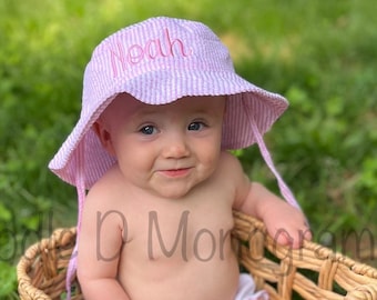 May include: A baby wearing a pink and white striped sun hat with the name "Noah" embroidered on it. The baby is sitting in a woven wicker basket.