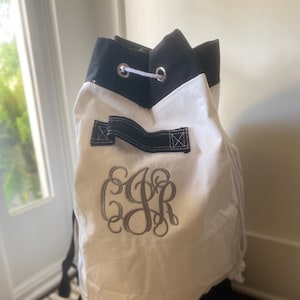 May include: A white and black drawstring bag with a monogram. The bag has a black base and top trim, with a white body. The monogram is embroidered in gray thread. The bag has black straps and silver grommets.