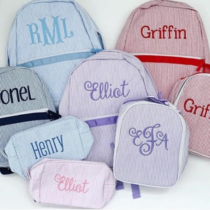May include: A collection of six personalized striped canvas backpacks and lunch bags in various colors. The bags feature embroidered names, including "Leonel", "RML", "Elliot", "Griffin", "Henry", and "EJA".