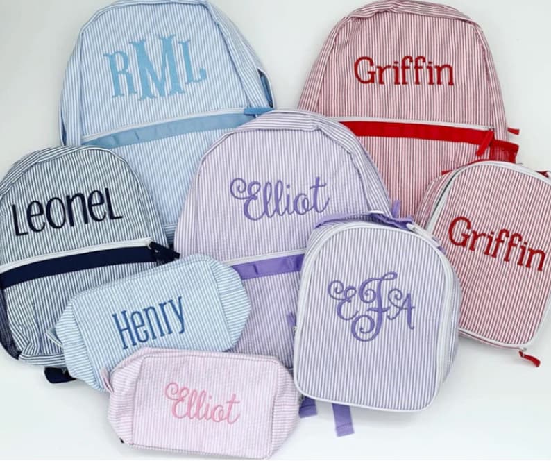 May include: A collection of six personalized striped canvas backpacks and lunch bags in various colors. The bags feature embroidered names, including "Leonel", "RML", "Elliot", "Griffin", "Henry", and "EJA".