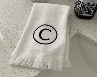 May include: White hand towel with a black embroidered letter C in a circle. The towel has a white fringe border.